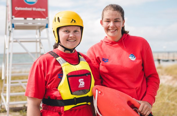 Zwei Rettungsschwimmerinnen der DLRG vor einem DLRG Wachturm. Sie lächeln in die Kamera.
